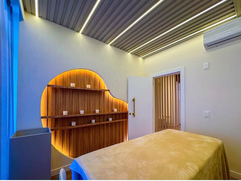 Sala de terapias com maca coberta por manta, nicho orgânico em madeira iluminado com velas e forro ripado com luz linear